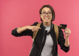 smiling young student girl wearing glasses back bag holding pointing credit card isolated pink wall scaled 1